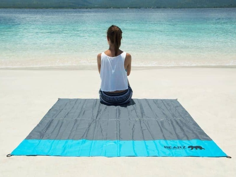 A feminine person sitting on a big, blue outdoor blanket on a sandy beach