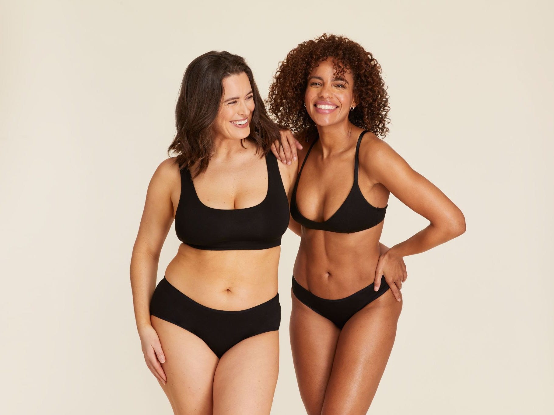 Two feminine persons smiling while modeling Andie swimsuits, specifically black two-pieces.